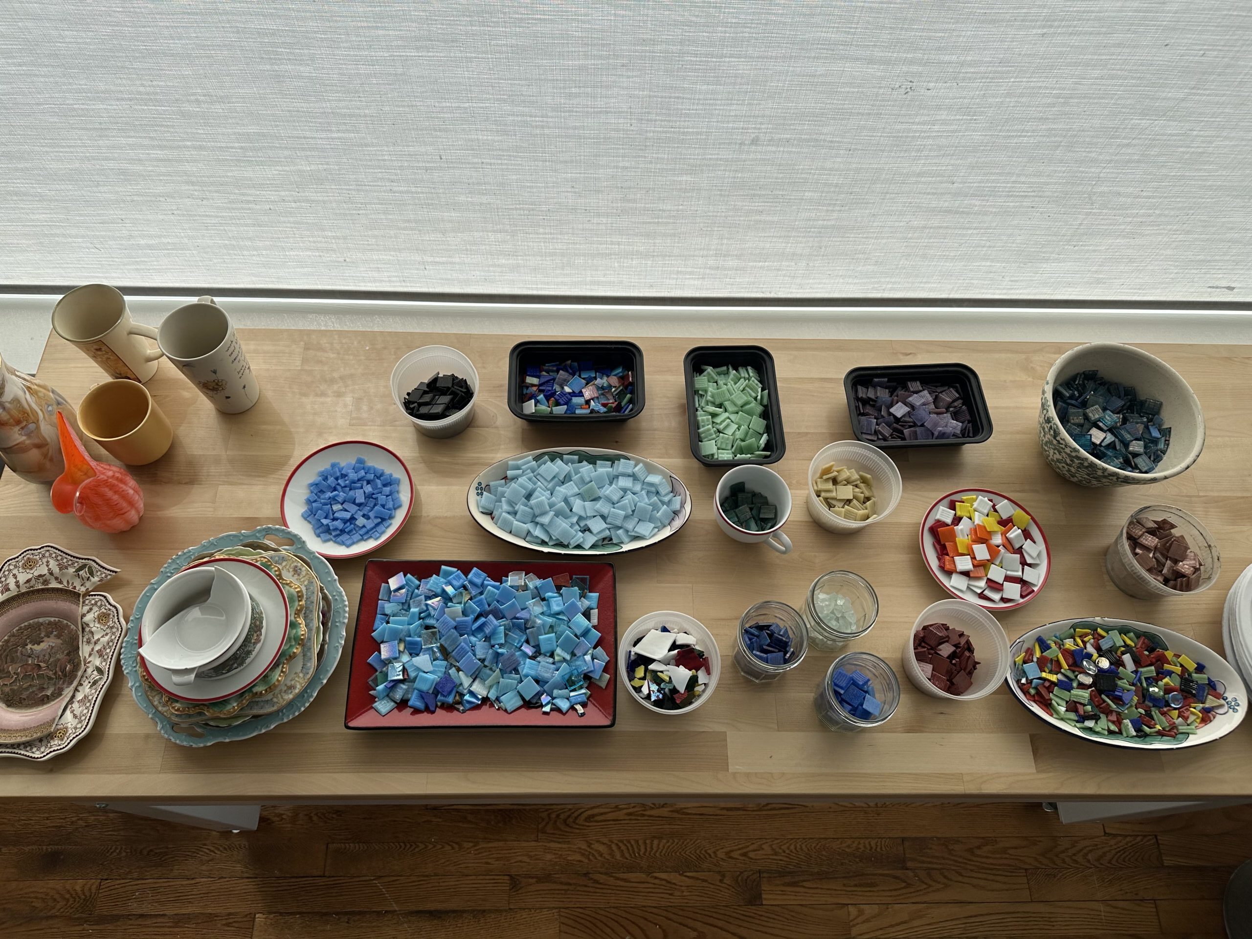 Overhead of table with assortment of jars, dishes, and vessels, many of which are filled with colorful glass tiles for mosaic.