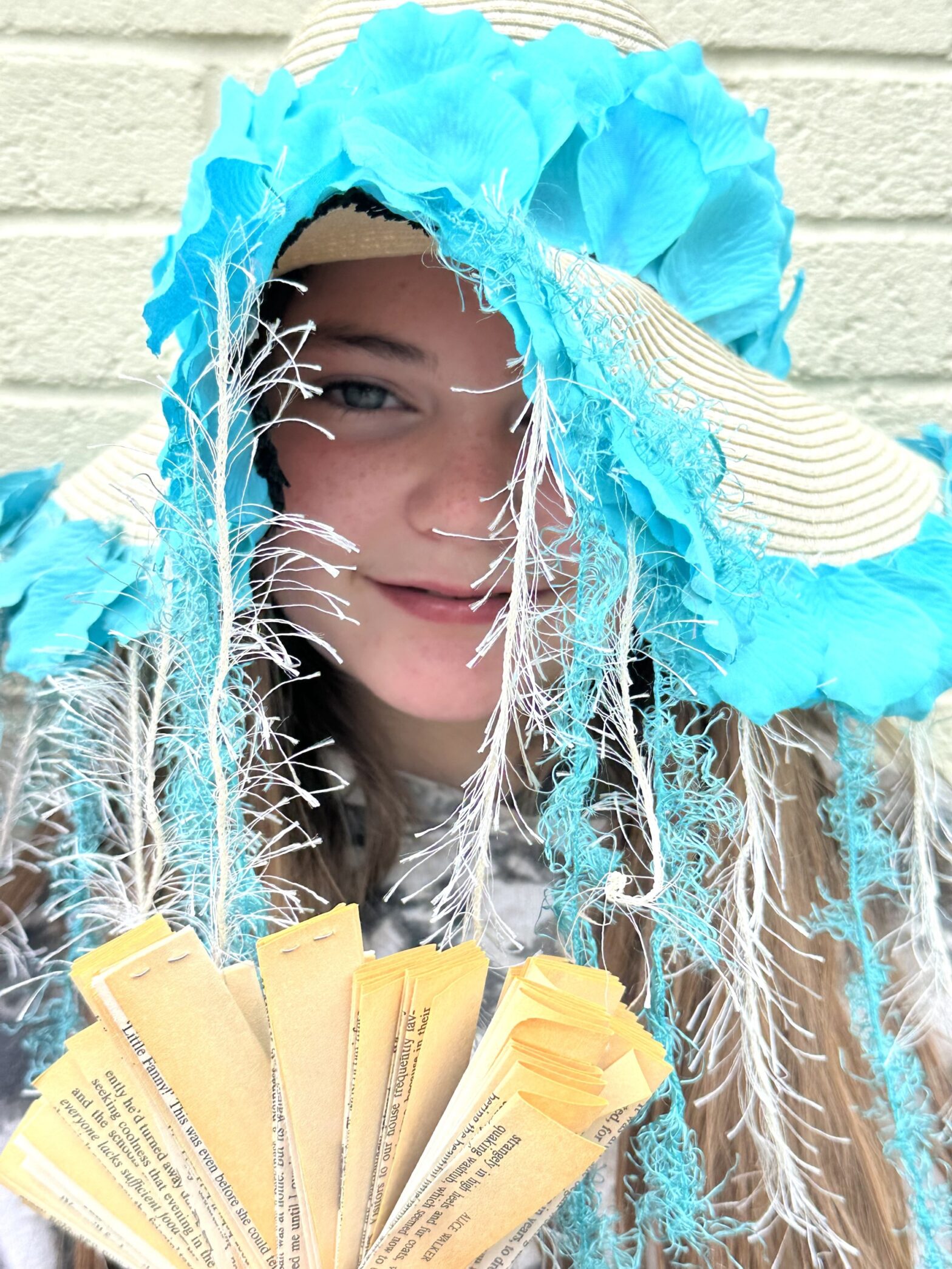 closeup of student wearing a hat made with turquoise fake flower petals and ribbon. she is smiling and holding lots of book pages folded and fanned out